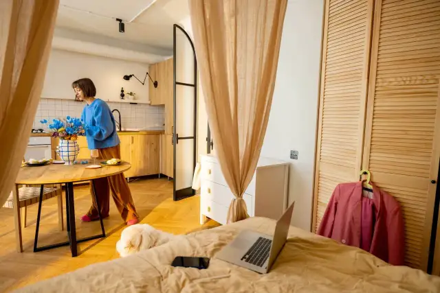 Kobieta w niebieskim swetrze aranżuje kwiaty w wazonie przy okrągłym stole. W kawalerce, mimo niewielkiej przestrzeni, wszystko jest na swoim miejscu.