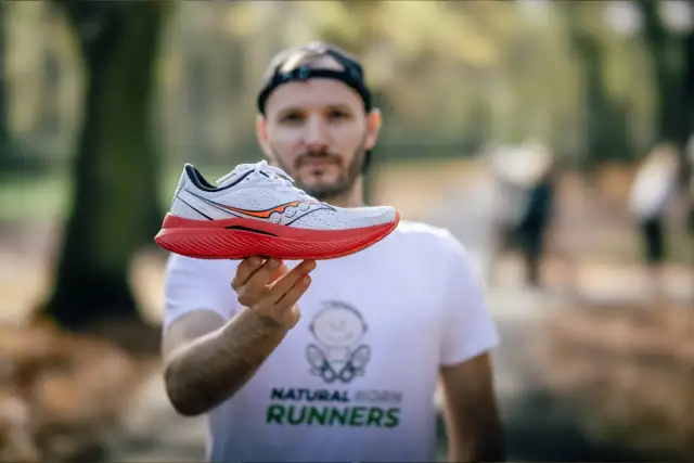 Mężczyzna w koszulce "Natural Born Runners" prezentuje biało-czerwony but biegowy Saucony. Saucony to znana firma z tradycjami.