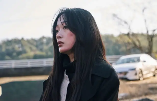 Młoda kobieta z zaczerwienionymi policzkami, wpatrzona w dal, gdy inni stali z boku. W tle widać samochód i most.