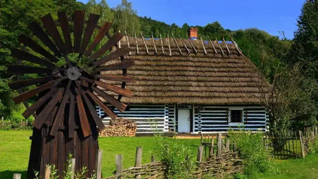 Drewniany wiatrak i tradycyjny dom z krytym strzechą dachem na tle zieleni.