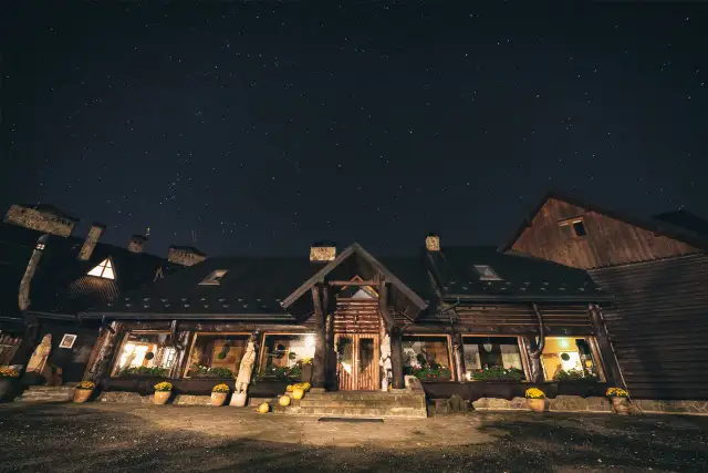 Nocne niebo pełne gwiazd nad drewnianą bacówką Biały Jeleń. Ciepłe światło sączy się z okien, zapraszając do środka.