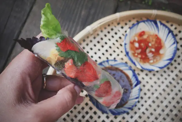 Ręka trzyma świeże rolls z papieru ryżowego z truskawkami i ziołami. Obok miseczki z sosem.