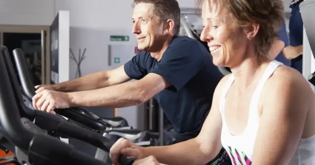 Uśmiechnięci ludzie ćwiczą na rowerach stacjonarnych. Czy ćwiczyć codziennie? Oni chyba tak robią, bo wyglądają na zadowolonych.