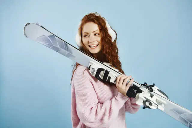 Uśmiechnięta rudowłosa dziewczyna w różowym swetrze trzyma narty. Dowiedz się, jak ustawić siłę wypięcia wiązań narciarskich dla bezpieczeństwa.