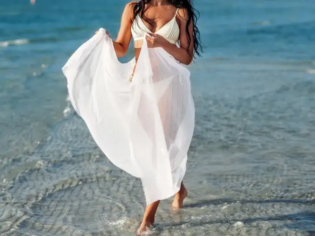 Kobieta w białym pareo spaceruje po plaży. To idealne okrycie na plażę, które chroni przed słońcem i dodaje elegancji.