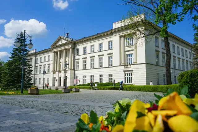 Biały budynek z kolumnami i zielenią, w tle błękitne niebo z chmurami.