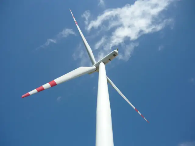 Biała mała elektrownia wiatrowa z czerwonymi pasami na łopatach obraca się na tle błękitnego nieba z chmurami.