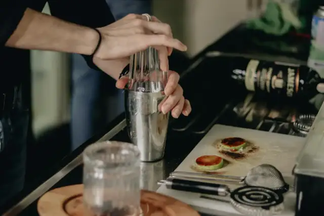 Ręce mieszają składniki w shakerze, przygotowując drinki na ciepło. Na blacie leży butelka whiskey i przypalone plasterki jabłka.