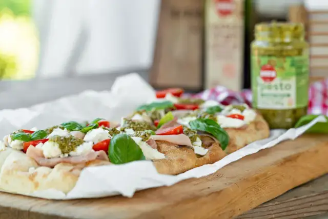 Długi placek z mozzarellą, pomidorkami i bazylią, polany zielonym pesto. Obok słoiczek z pesto.