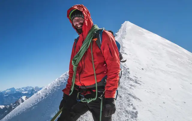Uśmiechnięty alpinista na szczycie Mont Blanc, w pomarańczowej kurtce i z liną, cieszy się widokiem Alp.