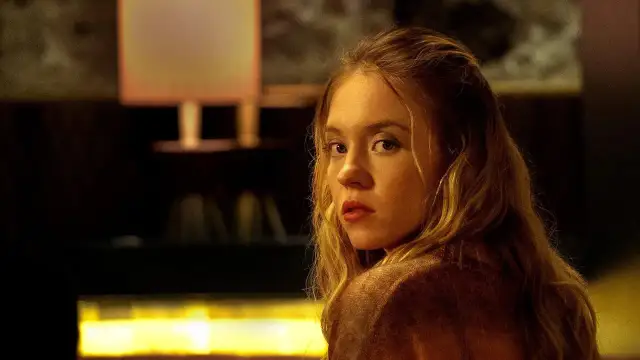 Młoda kobieta z burzą blond włosów spogląda przez ramię, jakby coś ją zaniepokoiło, kiedy nikt nie patrzy.