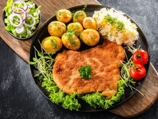 Talerz z panierowanym kotletem, ziemniakami, kapustą, surówką i pomidorkami.