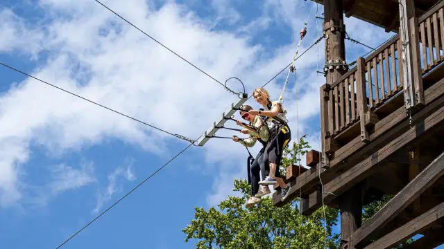 Dwie kobiety na tyrolce w parku linowym na tle błękitnego nieba z chmurami.