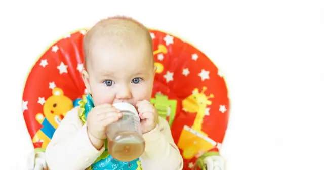 Niemowlę pije z butelki, siedząc w kolorowym krzesełku. Może to być uspokajająca melisa dla niemowlaka.