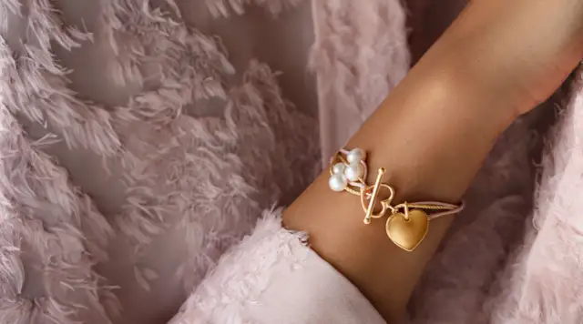 Złota bransoletka z sercem i perłami na różowej wstążce, na tle pudrowej tkaniny.