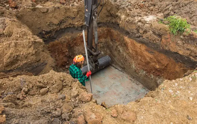 Koparka umieszcza rurę w wykopie. Pracownik w kasku i zielonej koszuli obserwuje, zastanawiając się, jaki kamień pod drenaż oczyszczalni będzie najlepszy.