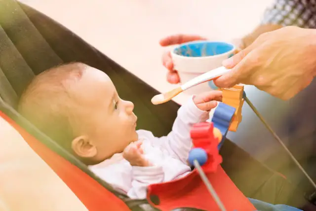 Karmienie 4-miesięcznego niemowlaka łyżeczką. Rodzic podaje jedzenie, dziecko patrzy z zaciekawieniem.