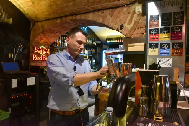 Mężczyzna nalewa piwo z kija w pubie, na ścianie wiszą etykiety piw z logo Mentzen.