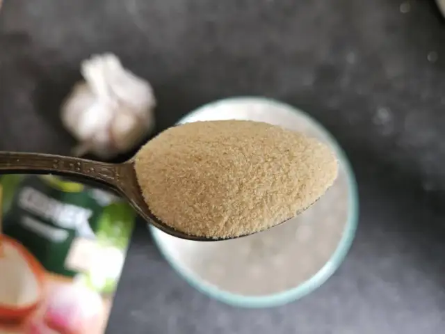 Łyżka z czosnkiem granulowanym na tle główki czosnku i opakowania.