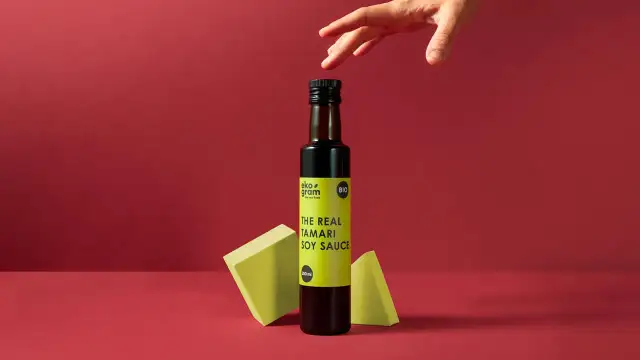 Butelka sosu tamari z bio-certyfikatem, na tle czerwieni, z ręką nad butelką.
