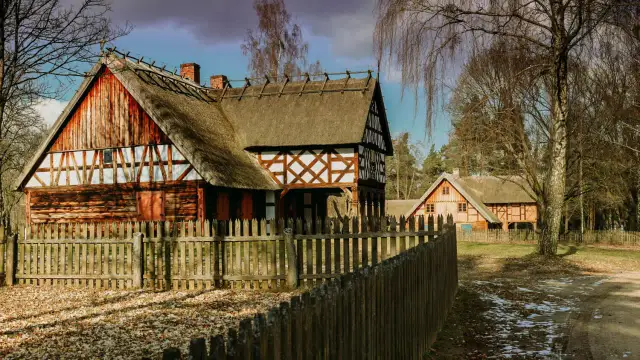 Drewniane domy z krytymi strzechą dachami i drewnianym płotem na tle drzew.