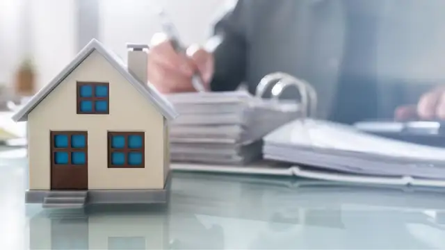 Model domu na biurku, obok stosy dokumentów i osoba pisząca. Czy kupując dom od dewelopera płacę podatek?