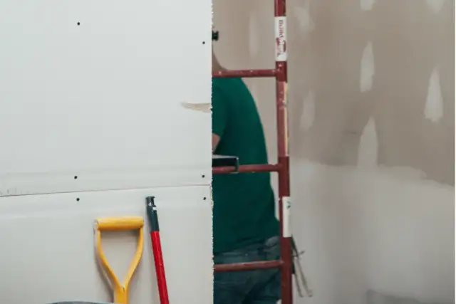 Prace remontowe w toku. Zastanawiasz się, ile schnie gips szybkoschnący? Na pierwszym planie łopata i trzonek, w tle pracownik budowlany.