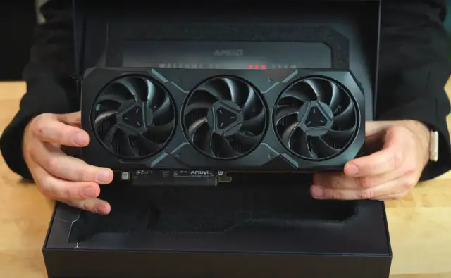 Ręce trzymają kartę graficzną Radeon RX 7900 XTX z trzema wentylatorami, wyjętą z czarnego pudełka z napisem "WELCOME TO RED TEAM".