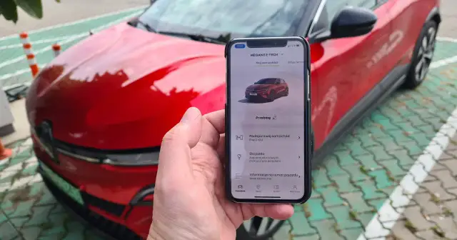 Aplikacja na telefonie pokazuje, jak przerobić auto na elektryczne, obok czerwonego samochodu.