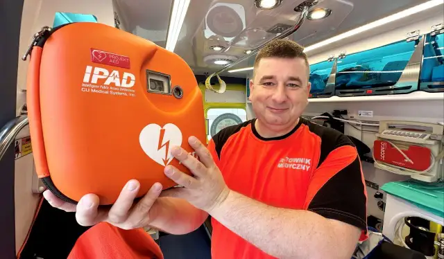 Ratownik medyczny trzyma pomarańczowy defibrylator AED w karetce.