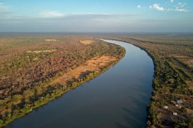 Szeroka rzeka płynie przez afrykański krajobraz. Po lewej stronie rozciąga się sucha sawanna, po prawej zielone drzewa i kilka domów.