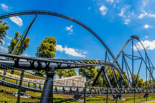 Tor roller coasteru Hyperion na tle błękitnego nieba z chmurami i zielonymi drzewami.