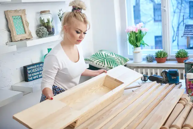 Kobieta składa drewniany stół, obok narzędzia i deski.