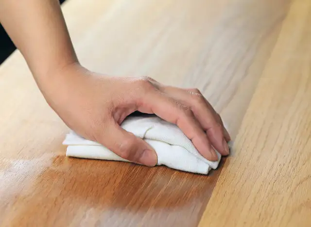 Dłoń z białą ściereczką poleruje drewniany blat, pokazując, jak odnowić stół lakierowany.