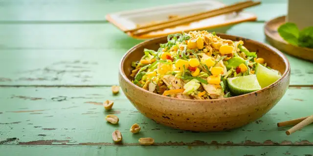 Kolorowa sałatka tajska z kurczakiem, kukurydzą, orzeszkami ziemnymi i limonką w glinianej misce na zielonym, drewnianym stole.