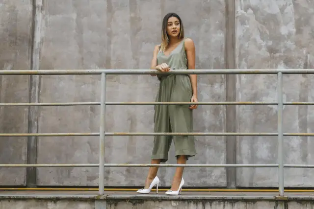 Kobieta w zielonym kombinezonie i białych szpilkach stoi przy metalowej balustradzie na tle betonowej ściany.