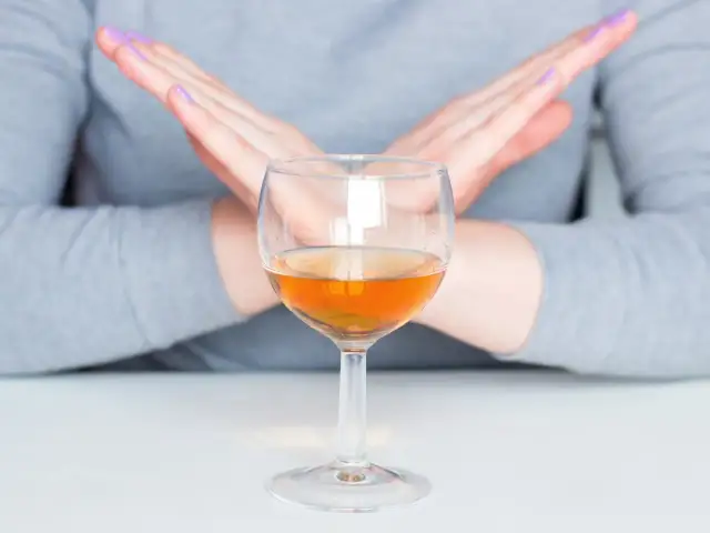 Dłonie skrzyżowane na znak stopu nad kieliszkiem z bursztynowym płynem. To symbol uczulenia na alkohol.