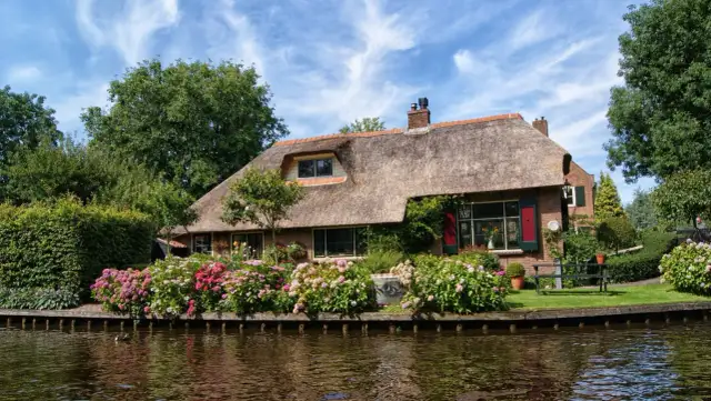 Uroczy domek z krytym strzechą dachem, otoczony kwitnącymi krzewami i wodą.