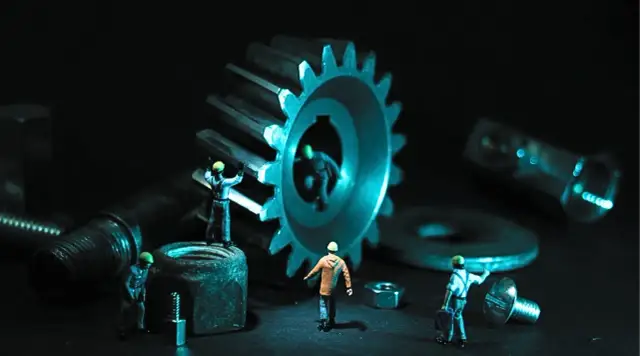Dział utrzymania ruchu pracuje nad wielką zębatką. Małe figurki robotników w kaskach porządkują śruby i nakrętki.