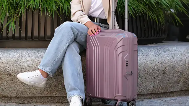 Różowa walizka z tworzywa sztucznego obok siedzącej osoby w jeansach i białych trampkach.
