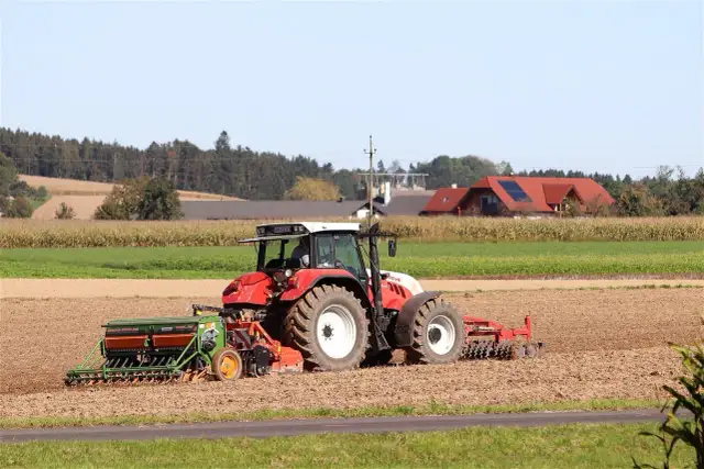 Czerwony traktor z siewnikiem na polu, w tle domy i las.