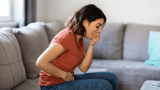 Kobieta siedzi na kanapie, trzymając się za brzuch i zakrywając usta, odczuwając mdłości.