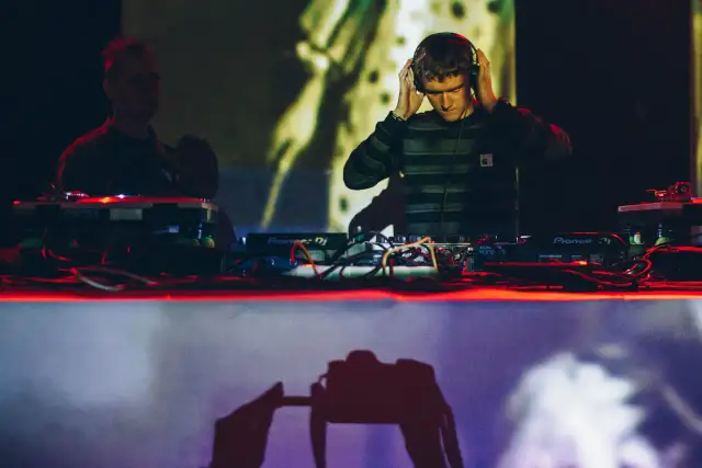 DJ w słuchawkach przy konsoli Pioneer DJ, w tle inny DJ i projekcja.