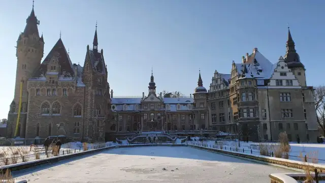 Zamek Moszna w zimowej scenerii, z zamarzniętym stawem i ośnieżonymi dachami. Bajkowa budowla prezentuje się majestatycznie.