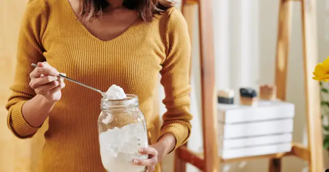 Kobieta nabiera łyżeczką sodę oczyszczoną do słoika. Czy soda oczyszczona jest zdrowa? W tle widać mydła i kwiat.