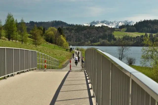 Rowerzyści na malowniczej trasie rowerowej w Czorsztynie, z widokiem na jezioro i ośnieżone szczyty Tatr.