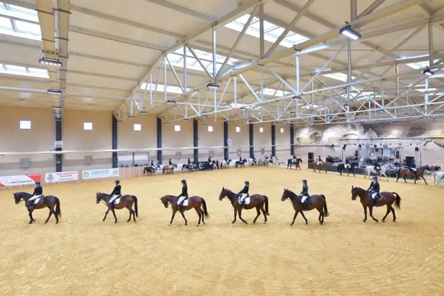Jeźdźcy w kaskach na koniach w hali ujeżdżalni.