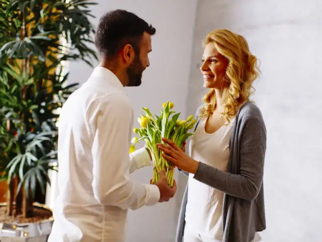 Mężczyzna wręcza kobiecie bukiet żółtych kwiatów. Uśmiechnięta kobieta z blond włosami przyjmuje prezent.
