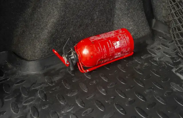 Czerwona gaśnica samochodowa leżąca na czarnej, gumowej macie w bagażniku.