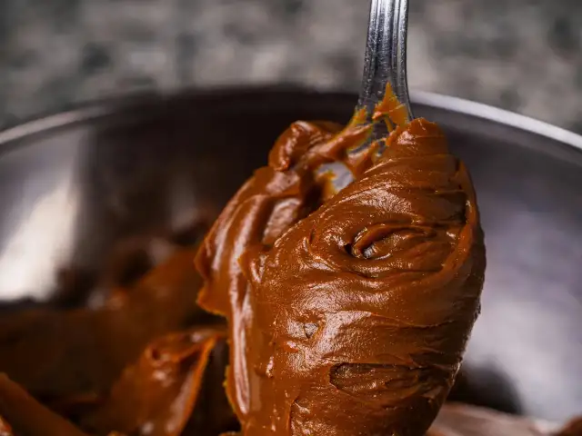 Gładka, brązowa masa czekoladowa na łyżce, w tle metalowa miska.